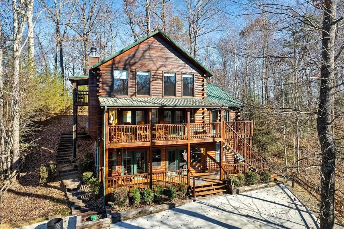 The image features the inviting exterior of a spacious log cabin nestled among tall trees in Sevierville, Tennessee. Expansive wooden decks wrap around the cabin, offering ample space for relaxing or dining outdoors. A stone pathway leads up to the entrance, suggesting easy access to nature walks and family gatherings. The setting is tranquil and surrounded by natural beauty, with the porch perfectly positioned for guests to watch sunsets or enjoy the fresh mountain air.