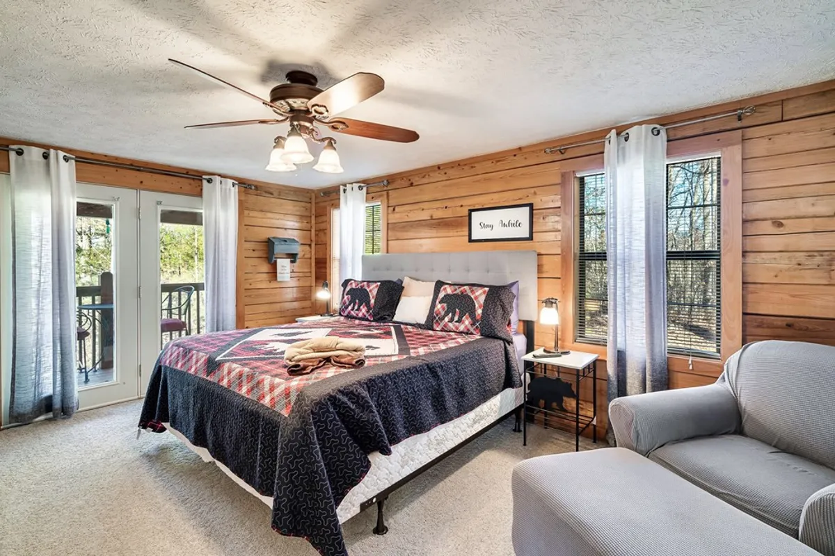 A serene bedroom featuring a bed with a patterned comforter, an inviting armchair by the window, and warm wooden paneling. Soft sheer curtains filter the natural light, and decorative wall art adds a touch of personality. The overall ambiance is calm and restful, perfect for relaxation after a busy day.