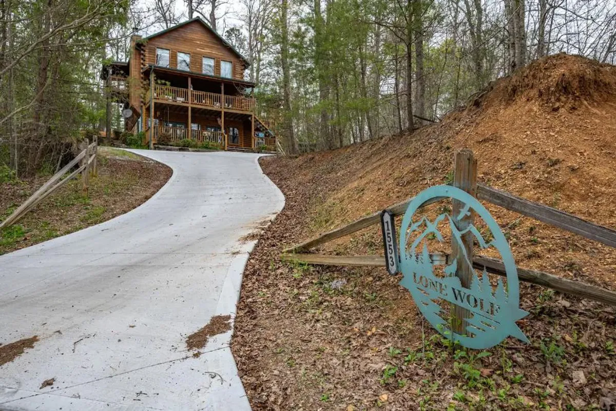 This image features the wooden cabin nestled within a serene forest setting, with a driveway leading up to the entrance and a 'Lone Wolf' sign visible. The surrounding trees and natural environment are prominent, emphasizing the cabin's proximity to nature and the outdoors.