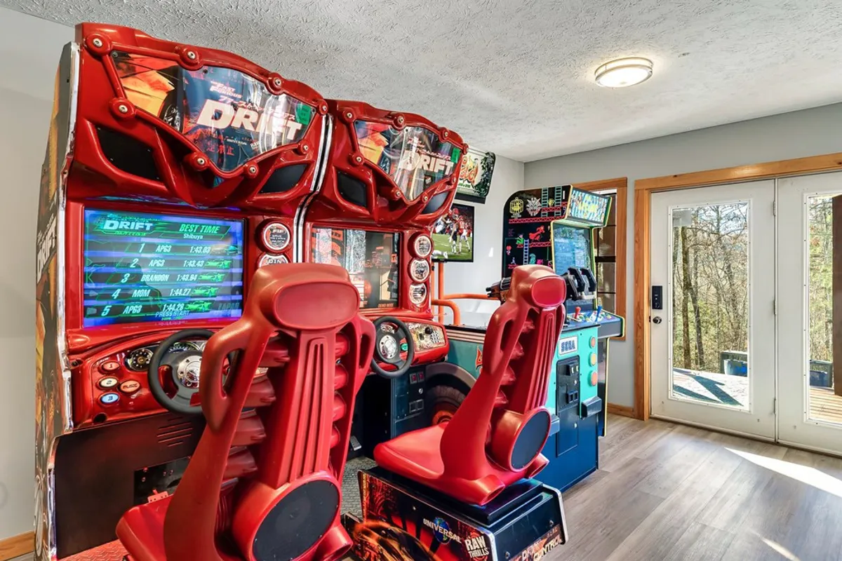 A vibrant arcade area featuring classic arcade machines with bright red racing seats and a view of the wooden deck entrance. The energy of the room is playful and nostalgic, inviting both kids and adults to join in friendly competitions.