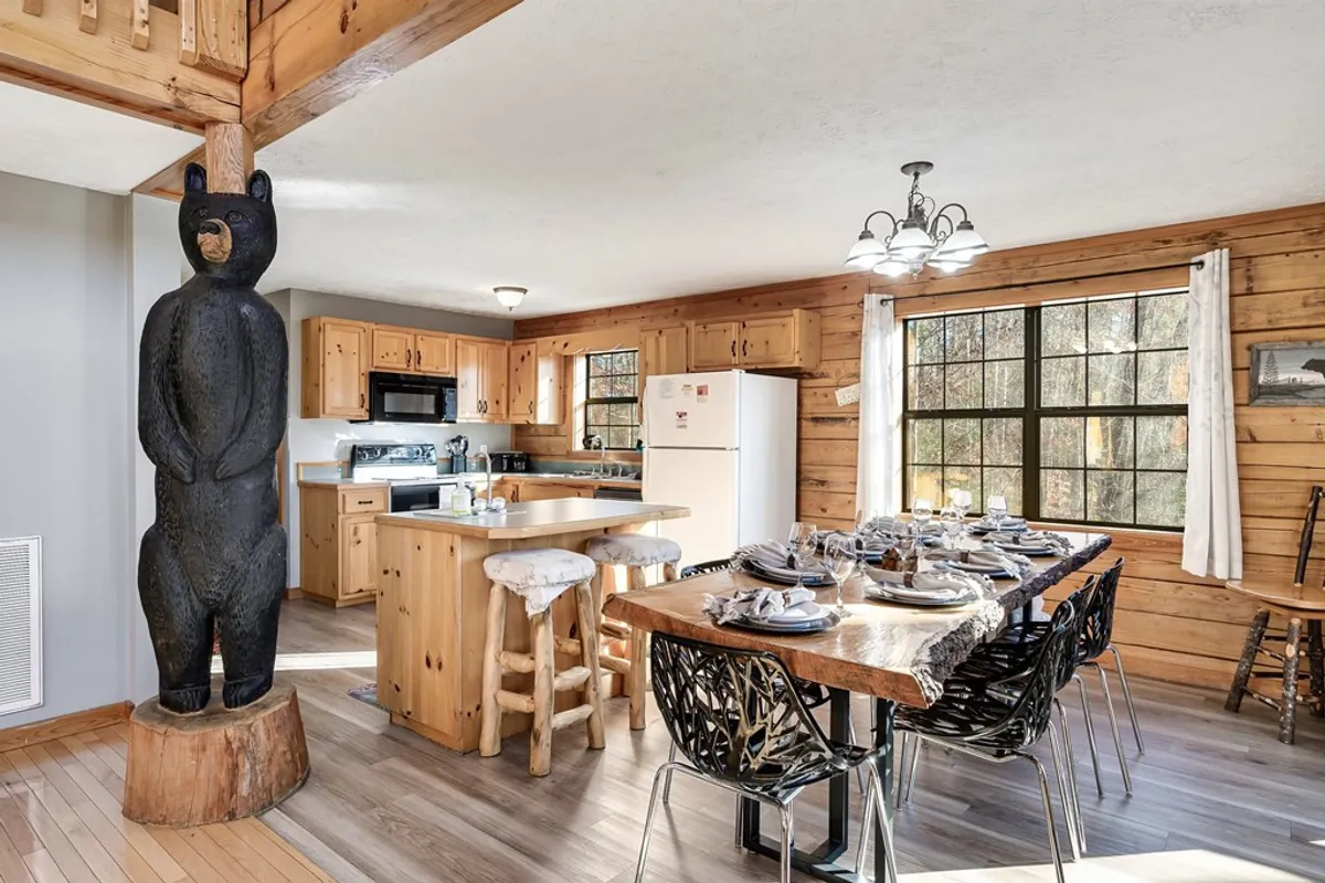 A charming view of the dining table set with place settings beside a kitchen island, watched over by a whimsical wooden bear statue. The arrangement blends the kitchen and dining spaces, suggesting shared family meals, playful storytelling, and collaborative cooking. The scene is bustling yet cozy, perfect for gatherings.