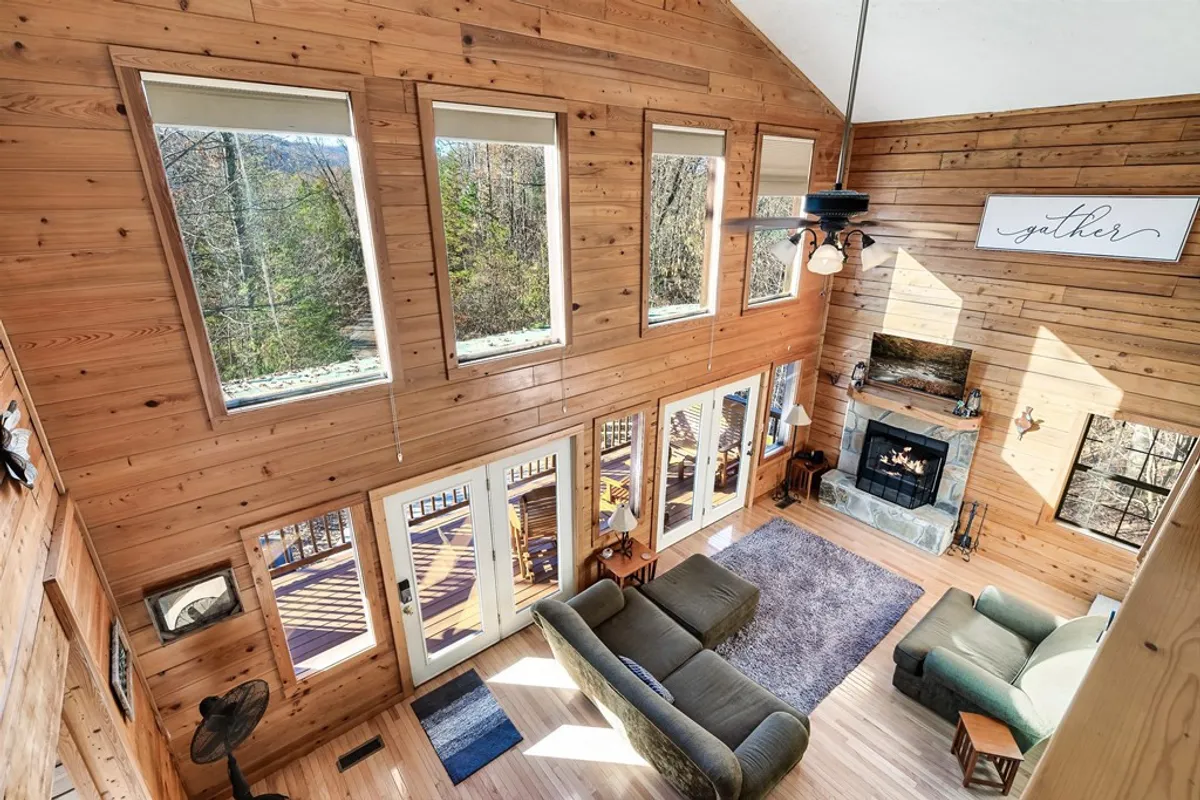 A warmly lit, open living area with a large, comfortable seating arrangement positioned in front of a striking stone fireplace. Tall windows frame the backdrop with natural light and woodland views, while a 'gather' sign above the fireplace subtly emphasizes the space’s purpose for togetherness. The wooden walls and ceiling beams add rustic charm and warmth, inviting relaxation and connection.