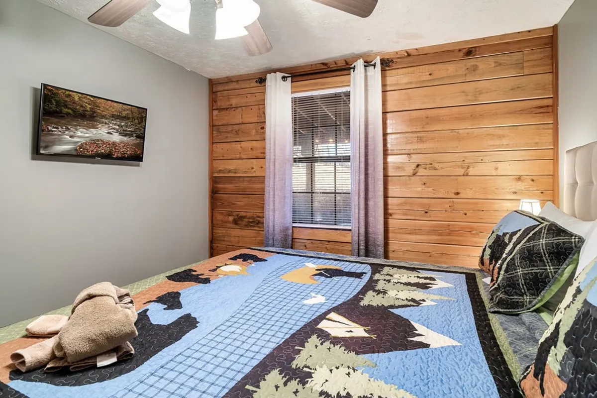 A warm, inviting bedroom with wooden wall panels, a colorful quilt on the bed, framed nature photography, and curtains that allow for privacy and natural light. The atmosphere is cozy and ideal for unwinding.