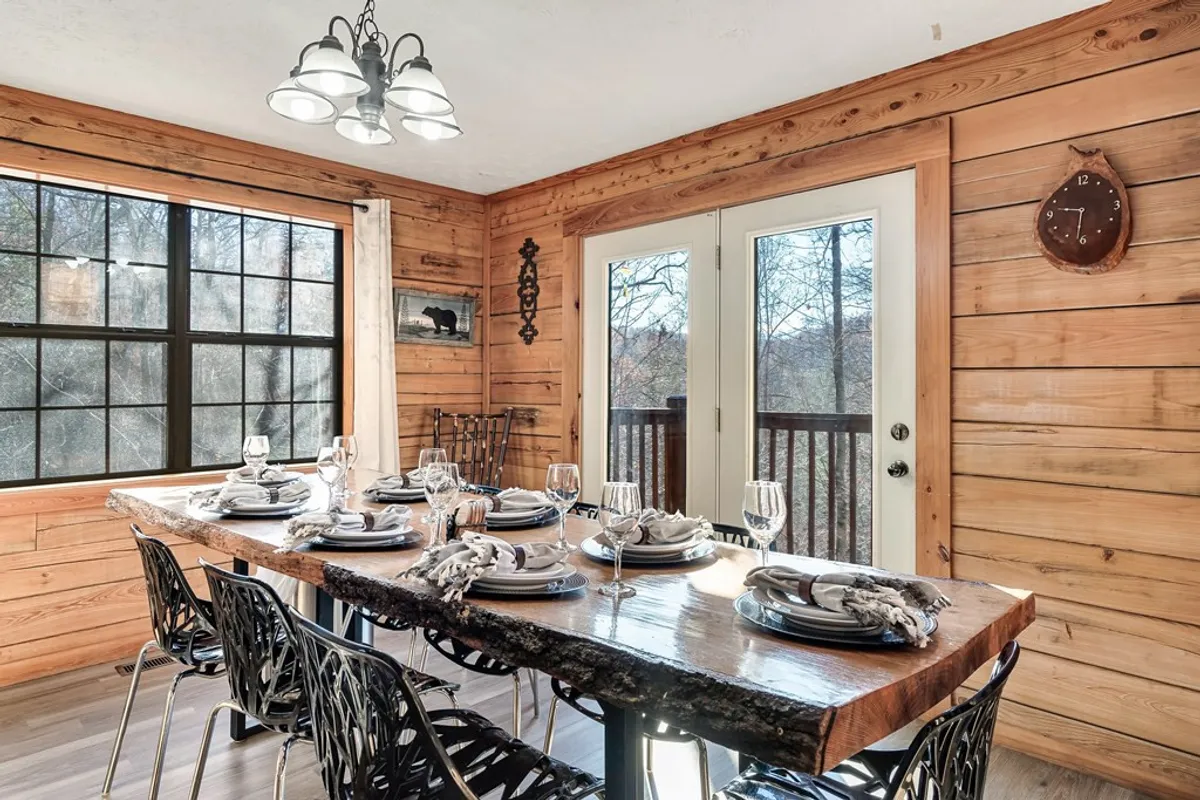 A spacious wooden dining area featuring a large table surrounded by black chairs, set for a shared meal. Double doors open to a balcony, bringing in natural light and offering a glimpse of the outdoors. Decorative touches like a wall clock and rustic accents evoke a sense of warmth and tradition, perfect for group dinners or celebratory feasts.